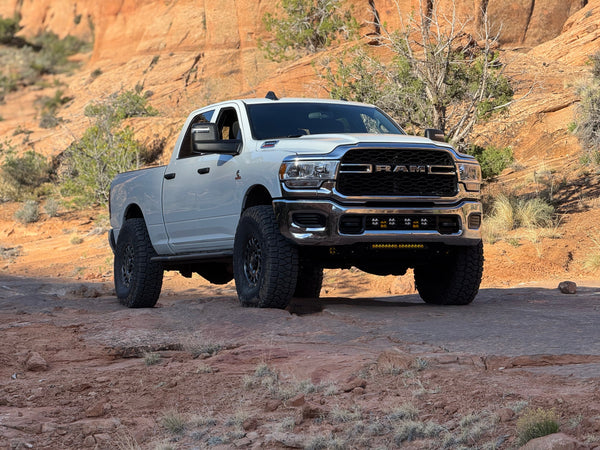 A white Ram HD crew cab on red slickrock, seen from the front, with a canyon wall behind it.