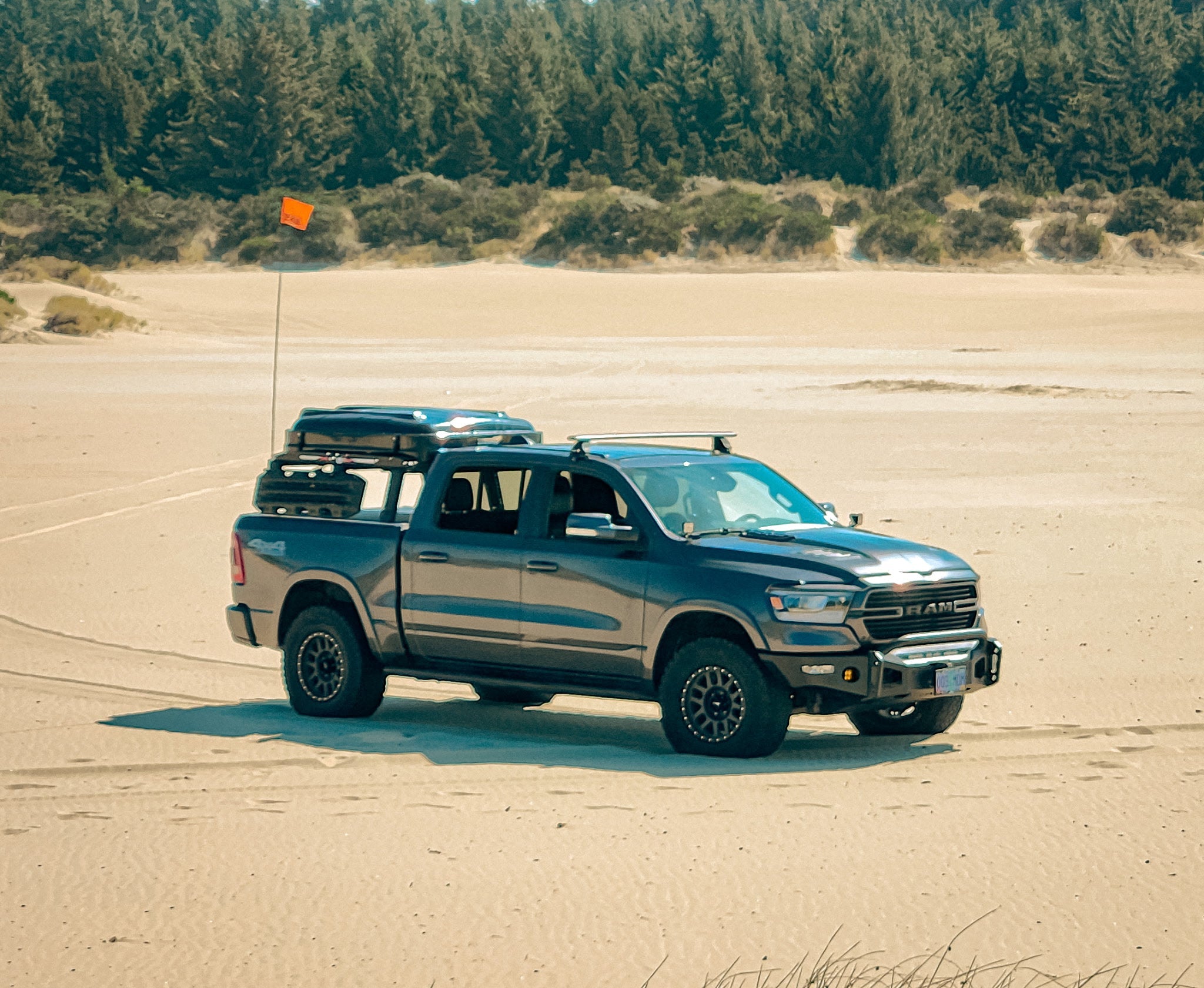 A grey Ram 1500 crew cab parked on sand, with a roof rack and cargo box and a pine treeline behind it.