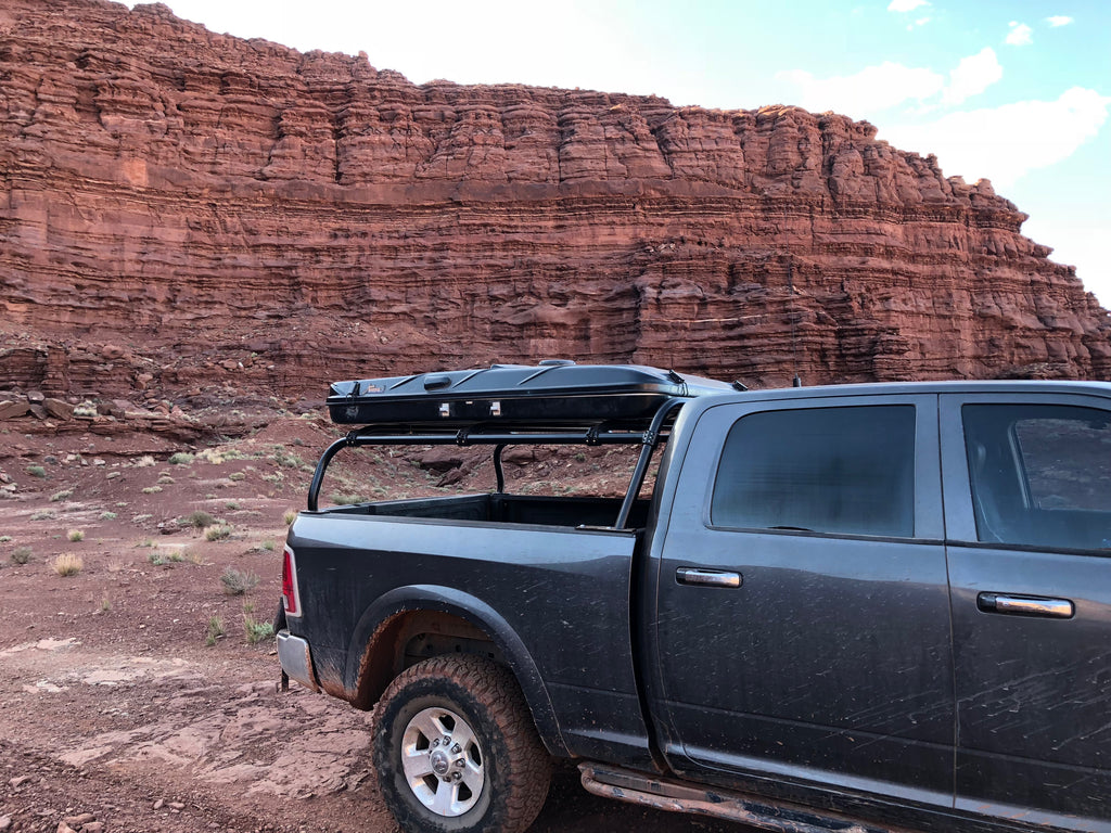 Side view of the grey Ram parked below a red rock wall, roof box on the rack