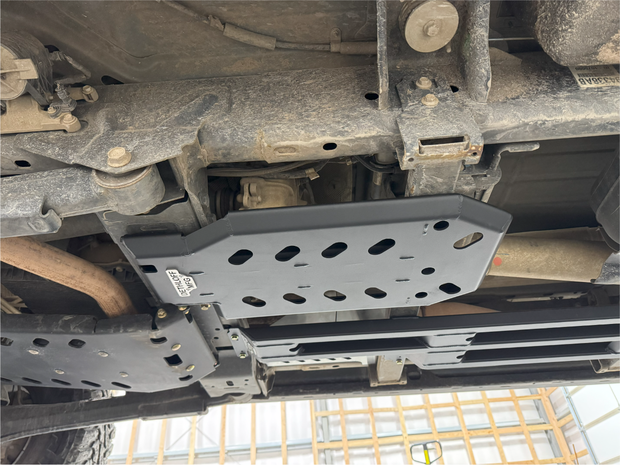 The guard in place under a truck on a lift, exhaust and driveshaft running alongside