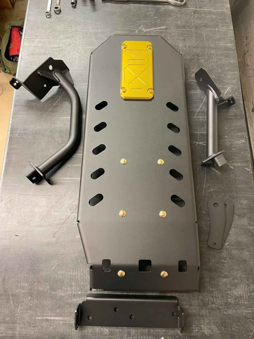 Skid plate on a steel workbench with its mounting brackets and tubular bar guards laid out beside it