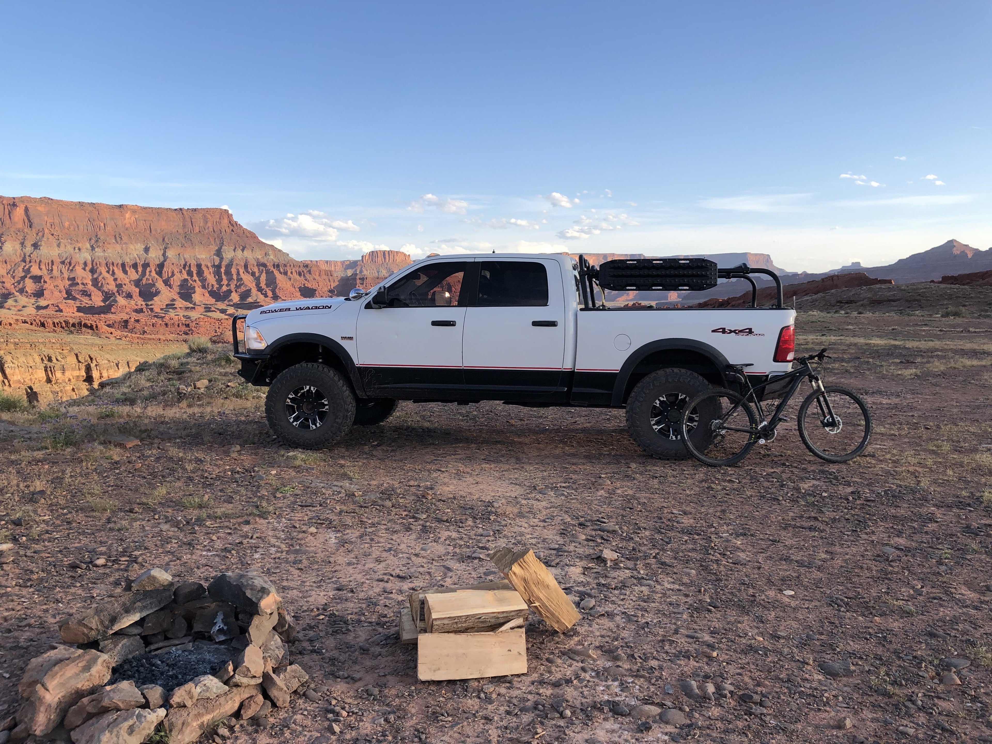 White Power Wagon parked at a canyon-rim camp, a bike leaning on the bed and a fire ring in the foreground