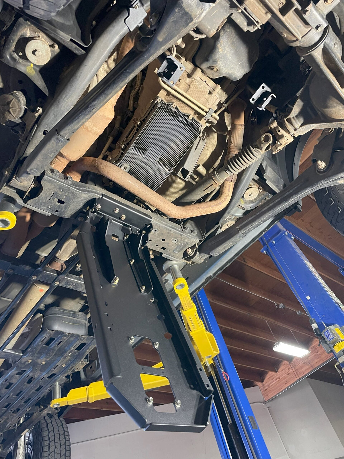 Truck raised on a two-post lift with the armor fitted, seen from below and behind