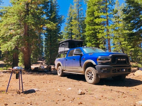 Blue Ram parked among pines at a rocky campsite, roof tent open on the bed rack