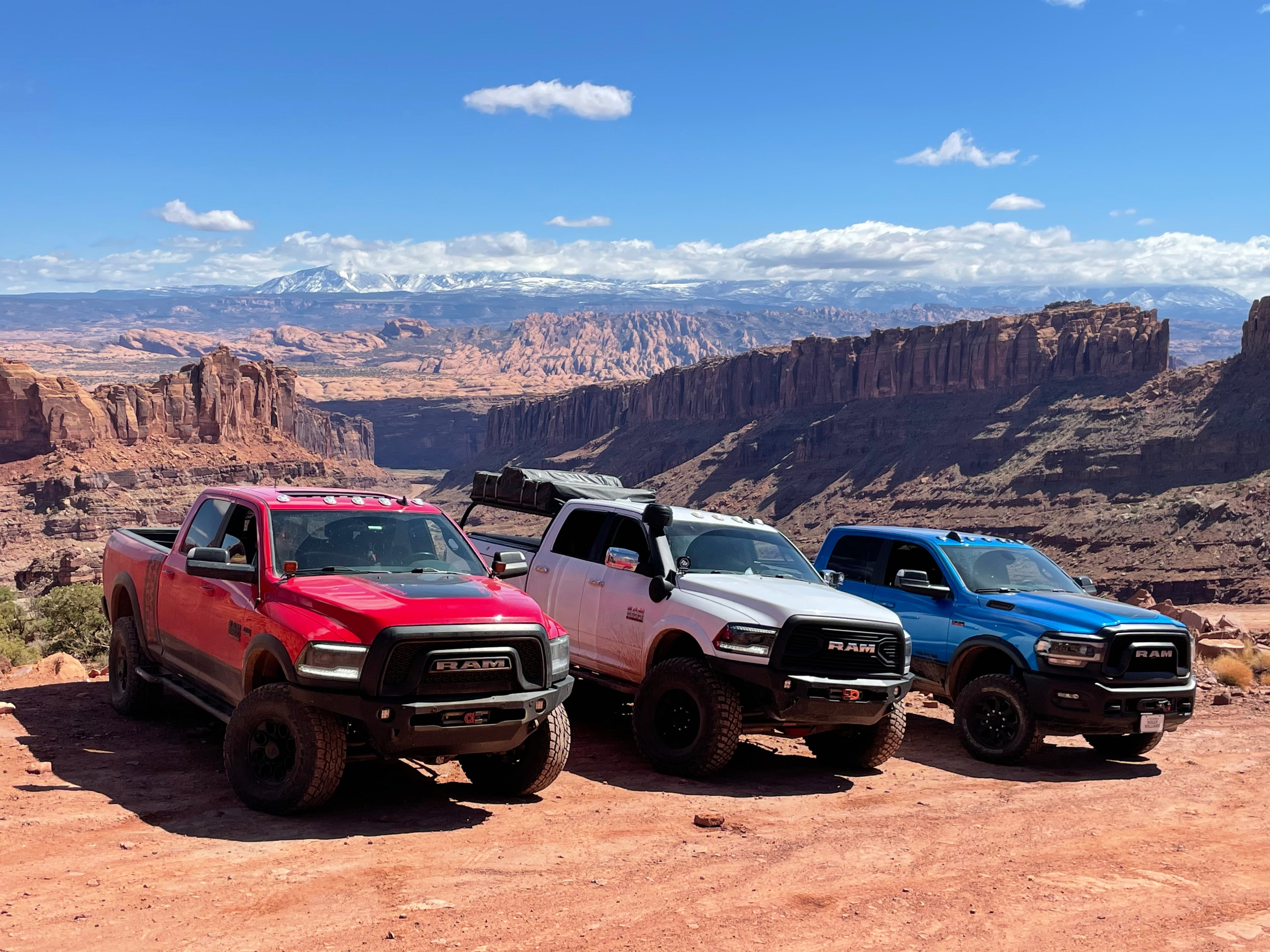 Three Ram heavy-duty trucks parked side by side on red slickrock at a canyon overlook, snow-capped mountains on the horizon.