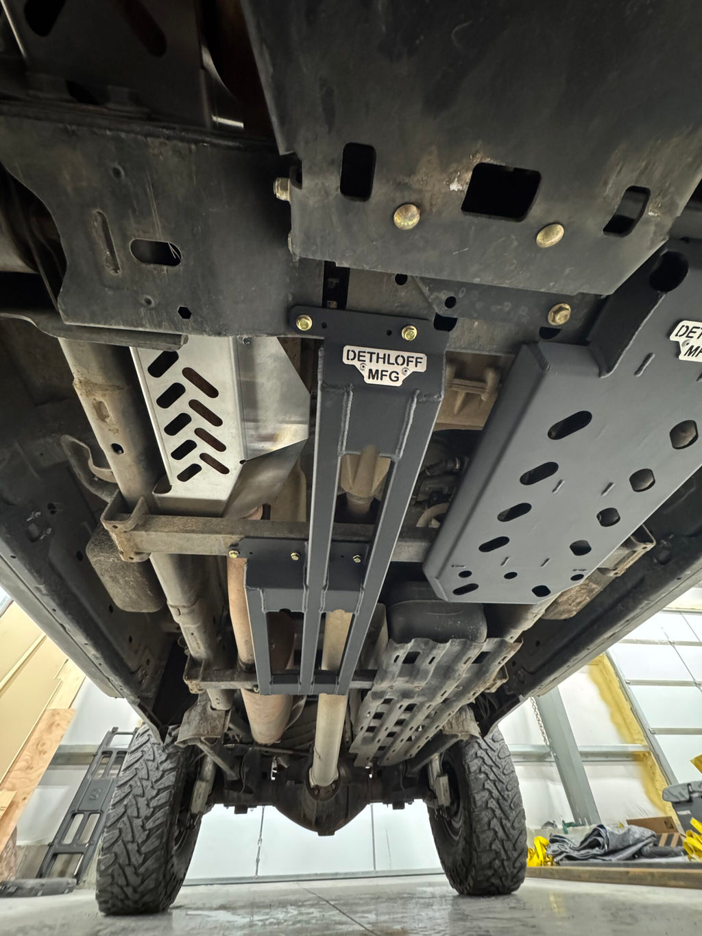 Underbody view with the DETHLOFF MFG badge on the centre brace and the slotted guard alongside the exhaust