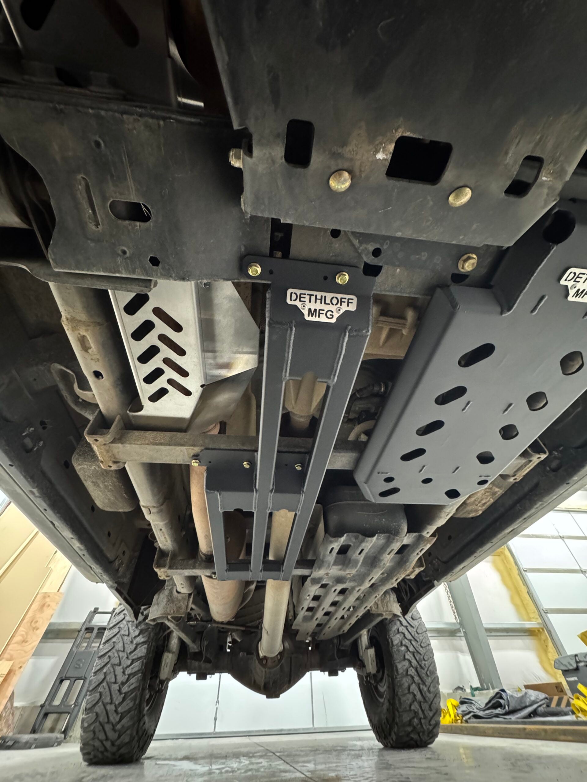 Underbody view with the DETHLOFF MFG badge on the centre brace and the slotted guard alongside the exhaust