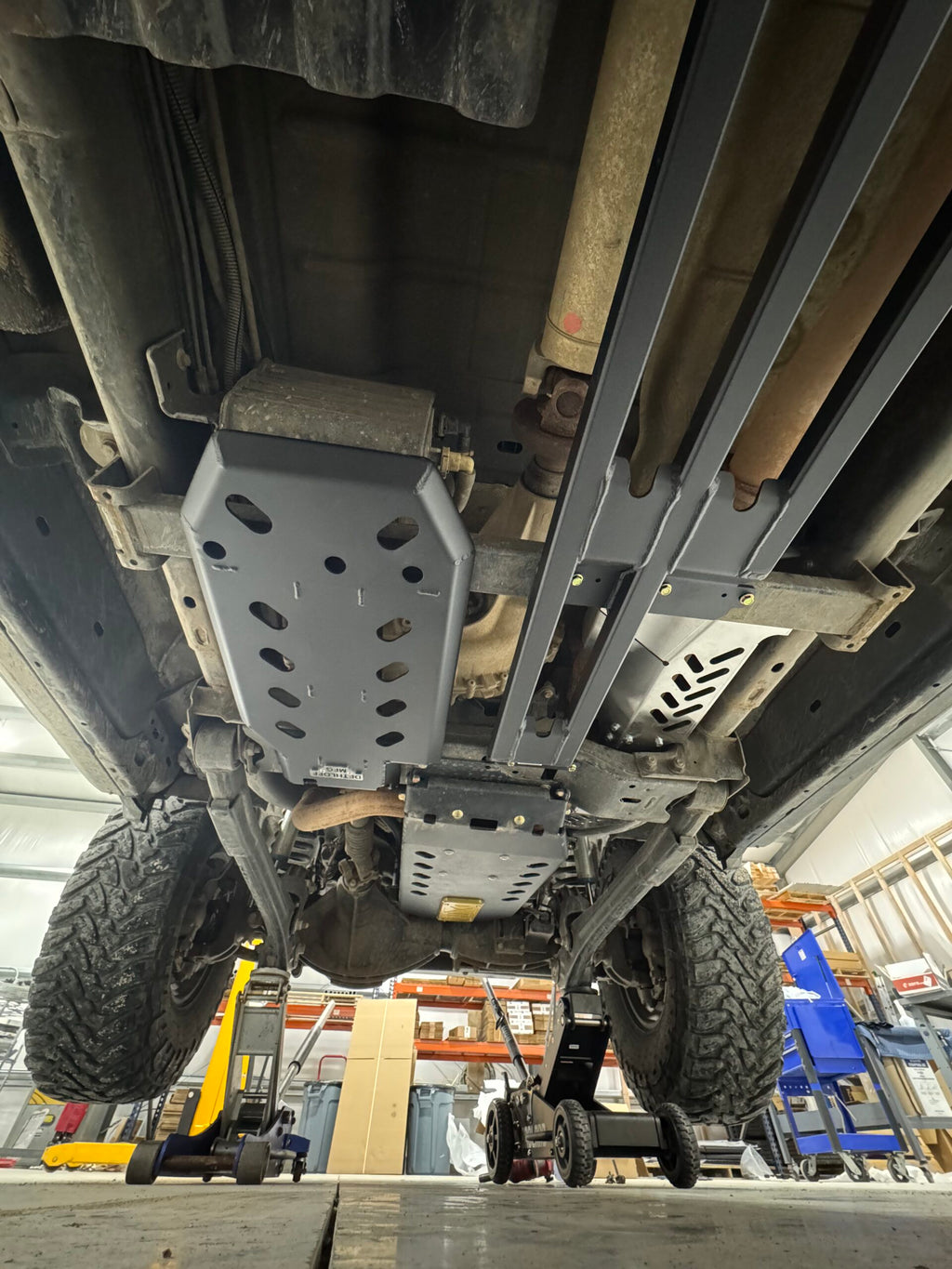 Looking straight up at the underbody on a lift, the bare aluminium guard sitting behind the grey Dethloff skid plates