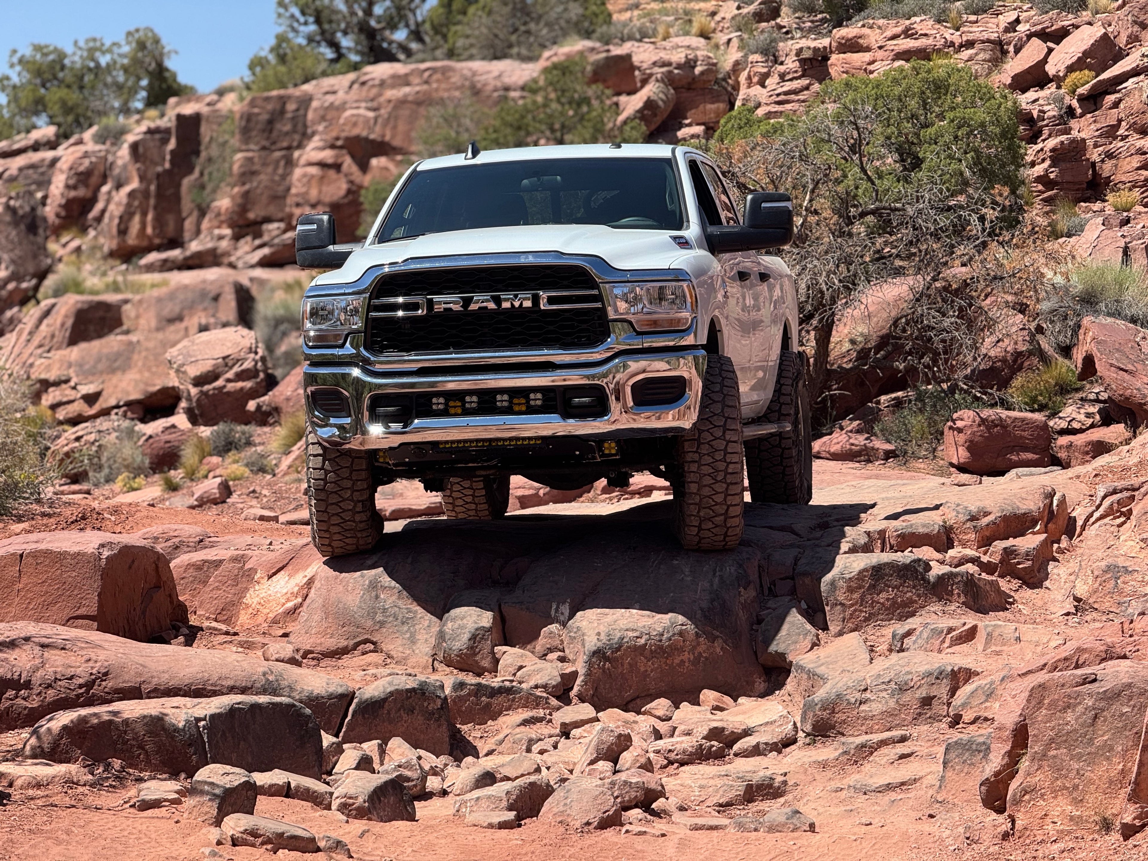White Ram HD climbing a red rock ledge, seen head on, with a front bumper carrying auxiliary lights.