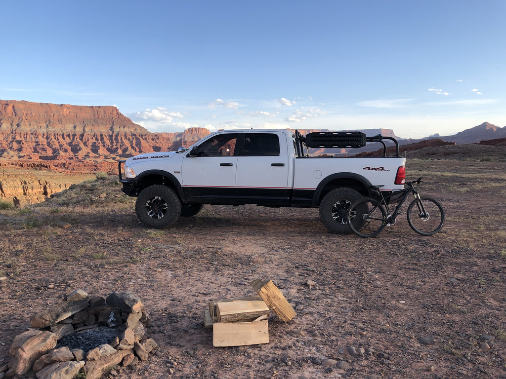 White Power Wagon parked at a canyon-rim camp, a bike leaning on the bed and a fire ring in the foreground