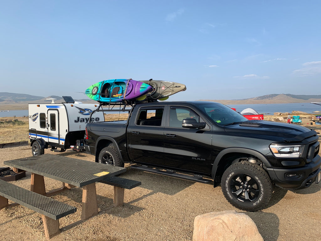Black Ram 1500 towing a camper trailer at a lakeside camp, kayaks loaded on the rack
