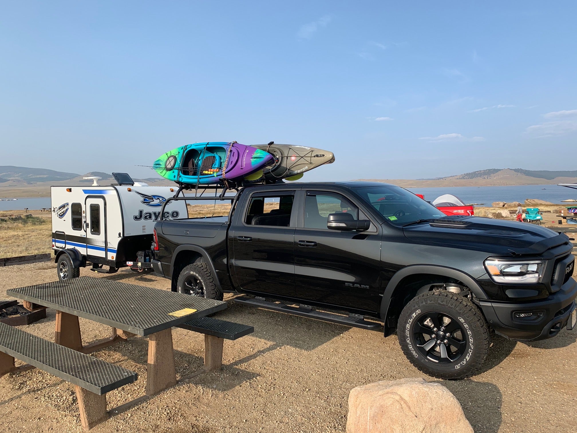 Black Ram 1500 towing a camper trailer at a lakeside camp, kayaks loaded on the rack